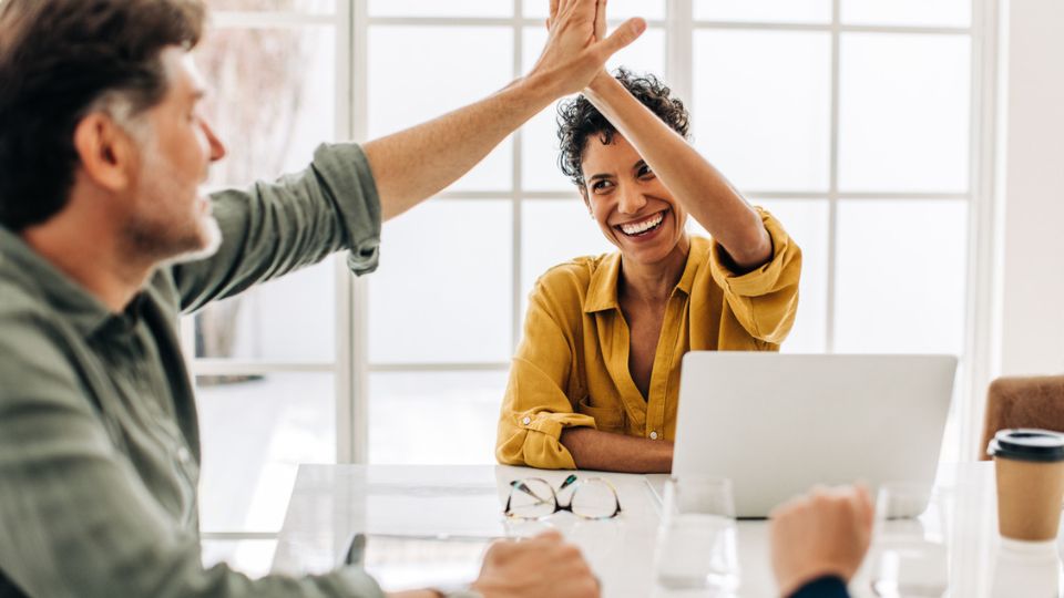 Lachende Frau sitzt im Büro am Laptop und gibt männlichem Kollegen ein High Five