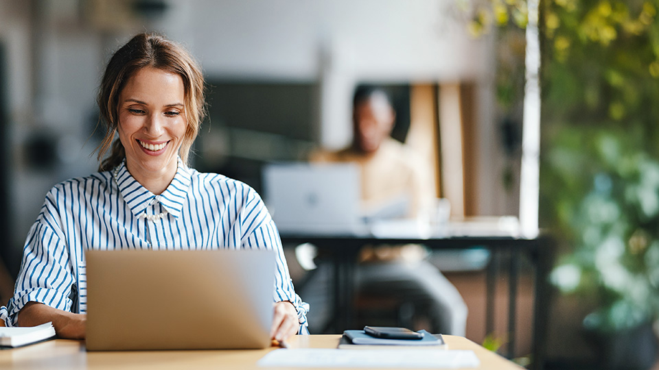 A smiling woman sits at a table, focused on her laptop